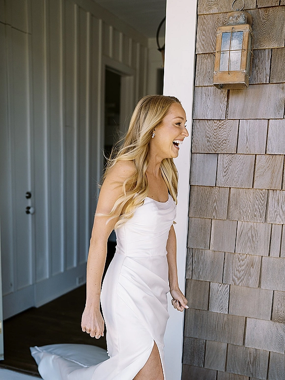 Bride portrait of a laughing bride in a strapless wedding dress with ruched bodice and slit, standing in a house doorway by a wall lantern