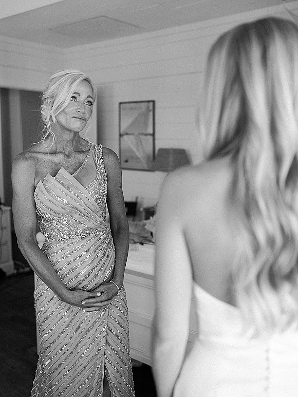 Wedding getting ready moment as mother of the bride in a beaded one-shoulder gown stands in a bedroom doorway, earrings sparkling