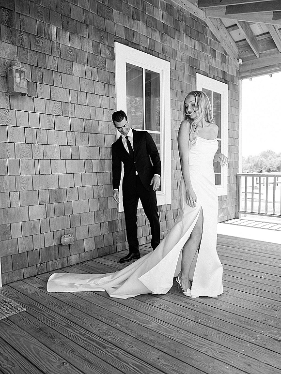 Couple portrait in a black and white wedding portrait, bride in strapless slit gown with train beside groom in black suit on shingle porch
