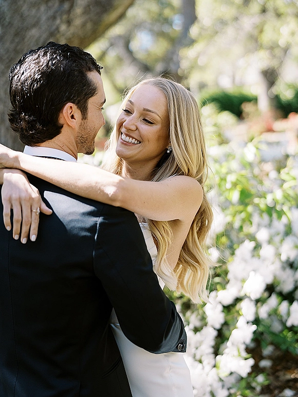 Couple portrait of bride and groom embrace in garden greenery, bride smiling at groom with a wedding ring accent in daylight