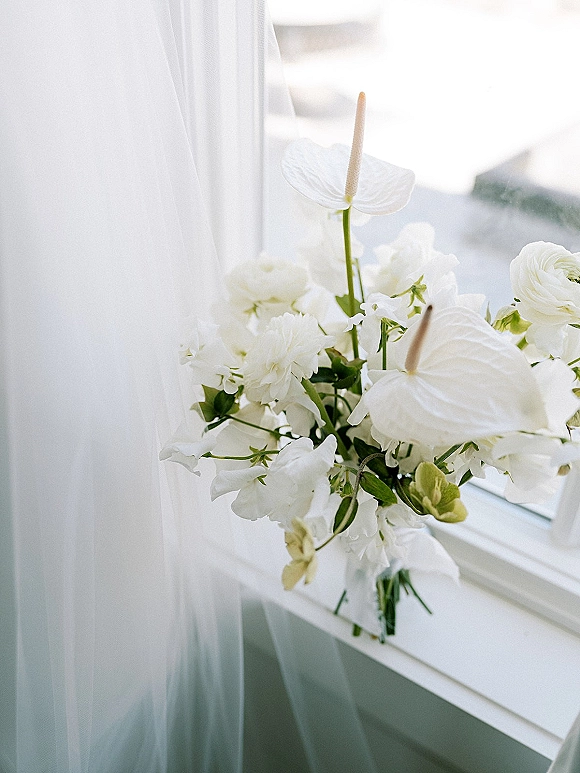 Bridal bouquet with white anthurium blooms and ranunculus wrapped in a sheer veil, resting on a windowsill in soft natural light
