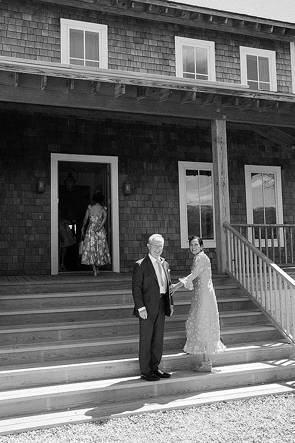 Couple portrait in a black and white wedding photo, bride in lace dress and heels holding groom’s hand on wooden porch steps
