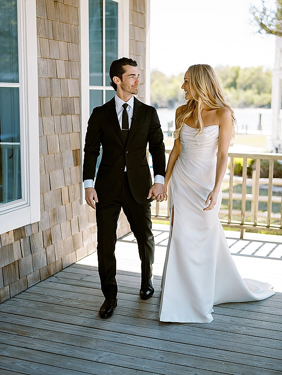 Couple portrait of bride and groom holding hands on a wood porch, strapless dress and black suit, with water and trees behind in daylight