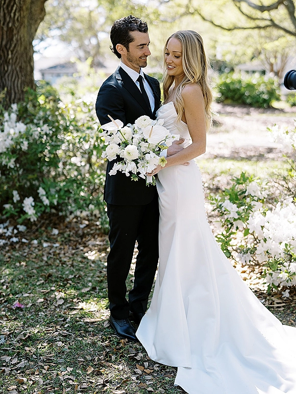 Couple portrait of bride in a strapless wedding dress holding a white bouquet beside groom in a black suit in a sunlit garden