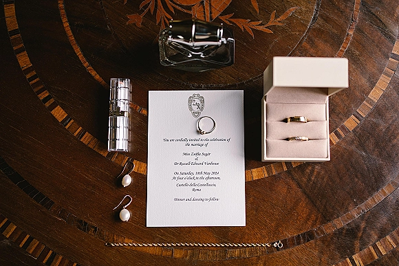 Invitation suite wedding invitation flat lay with monogram crest, rings, pearl earrings, perfume and lipstick arranged on an inlaid wood table
