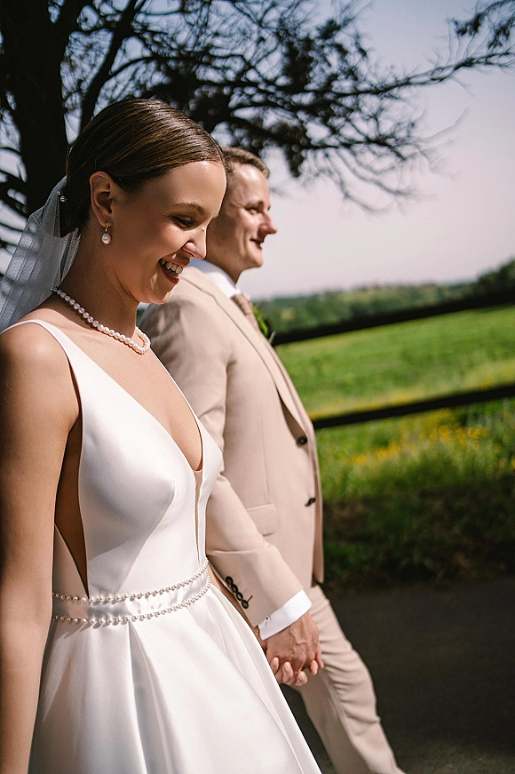 Couple portrait of bride and groom walking hand in hand, bride in veil with pearl jewelry, beside a rustic fence in green fields