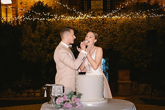 Wedding cake cutting as bride in pearl necklace and groom with boutonniere share a slice by string lights in a garden night setting