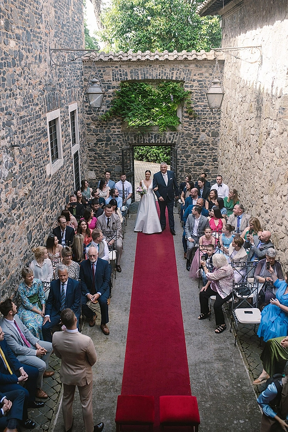Bridal processional with bride walking down aisle on a red runner, holding a bouquet in a stone courtyard with seated guests and lanterns