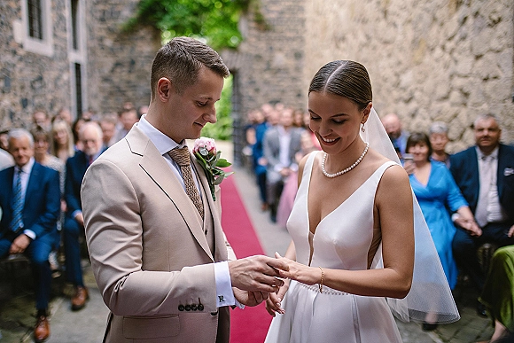 Wedding ring exchange as the groom places a band on the bride’s finger during an outdoor ceremony on a red aisle runner with guests and stone walls behind