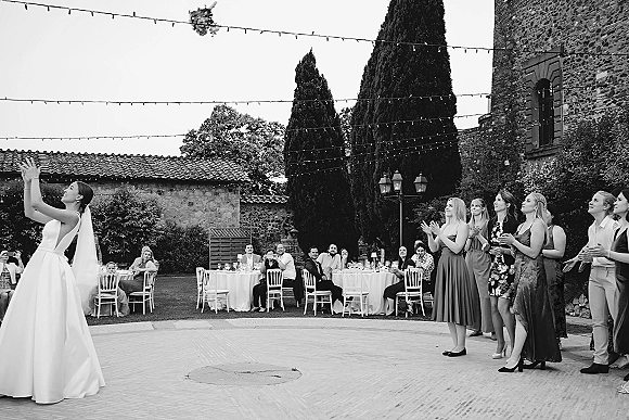 Bouquet toss as the bride throws a wedding bouquet in air, veil flowing under string lights in an outdoor stone courtyard reception
