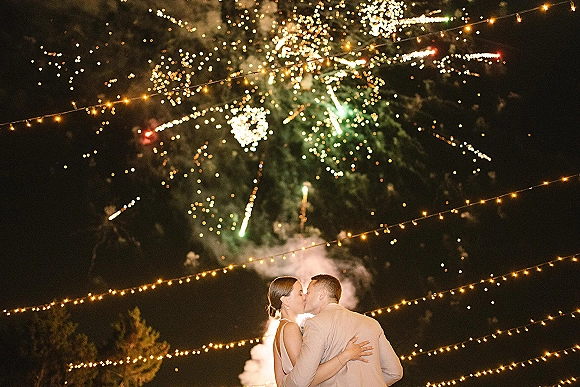 Wedding kiss beneath fireworks wedding exit, with string lights glowing overhead against a smoky night sky and dark trees