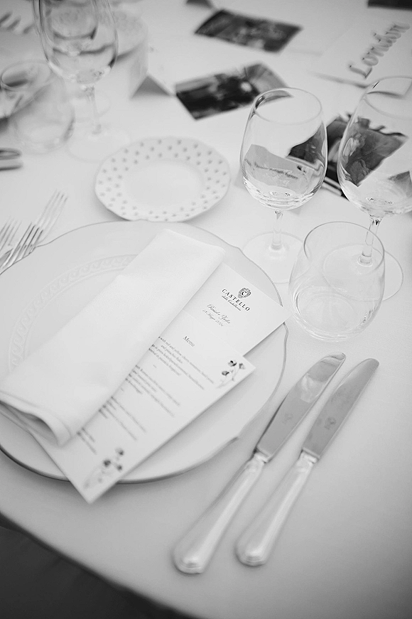 Wedding place setting with a wedding menu card on stacked plates, white napkin, silver flatware, and stemware on a white tablecloth