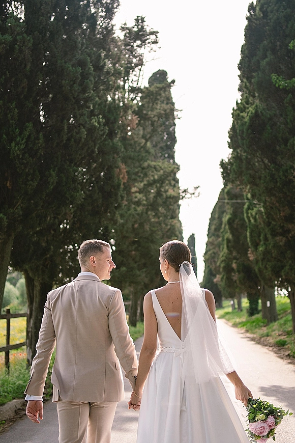 Couple portrait of newlyweds holding hands, walking away on a tree-lined road; bride’s long veil and bouquet beside groom in tan suit