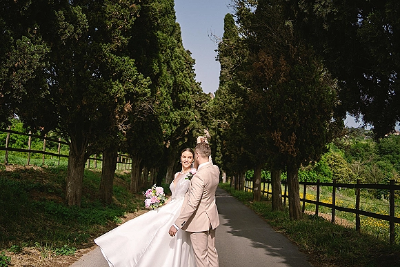 Couple portrait of bride and groom outdoors as she twirls her flowing gown with pink bouquet, walking a tree-lined road by a wooden fence