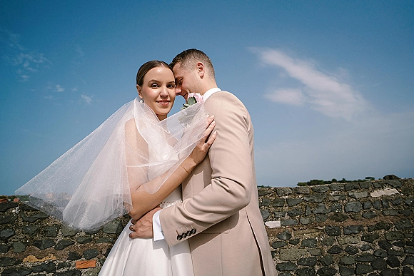 Couple portrait of bride and groom embrace as he kisses her cheek under a windswept veil by a stone wall and blue sky
