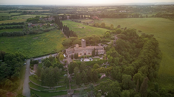 Wedding venue aerial view of a stone villa with round patio reception tables and string lights, framed by cypress trees at sunset