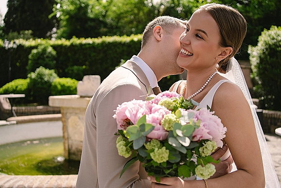 Wedding couple portrait with groom kissing bride’s cheek as she smiles holding a pink peony bouquet on a stone terrace by a fountain