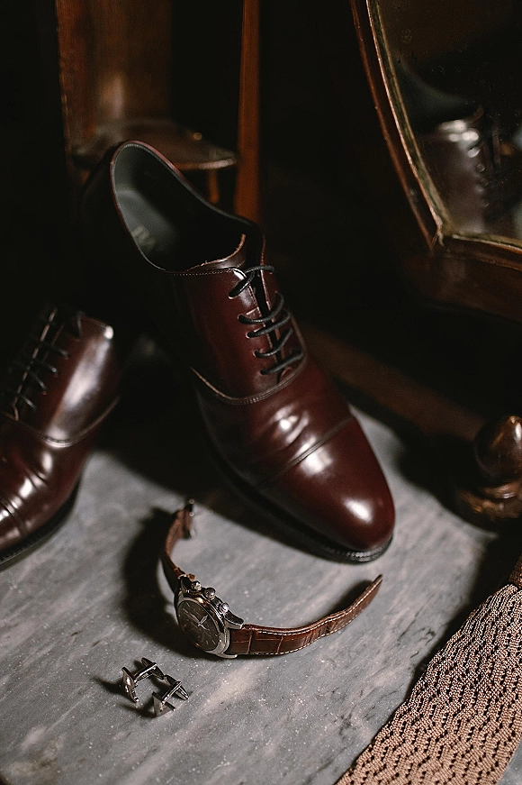 Groom accessories flat lay with brown leather dress shoes, wristwatch, cufflinks, and textured tie on marble by dark wood mirror