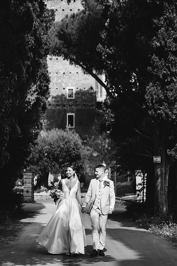 Wedding couple portrait of bride and groom walking hand in hand, her veil and bouquet flowing, on a tree-lined road by a stone estate building