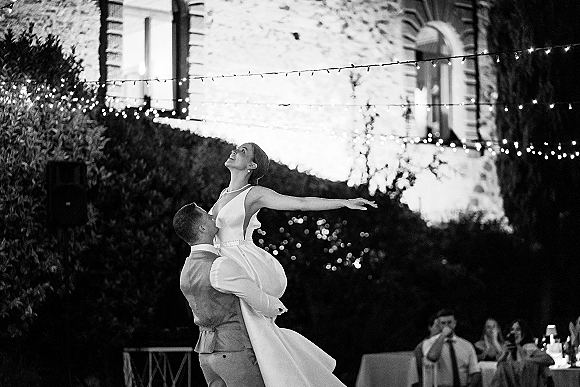 First dance as groom lifts bride in a wedding lift dance, her pearl necklace catching string lights in an outdoor garden reception