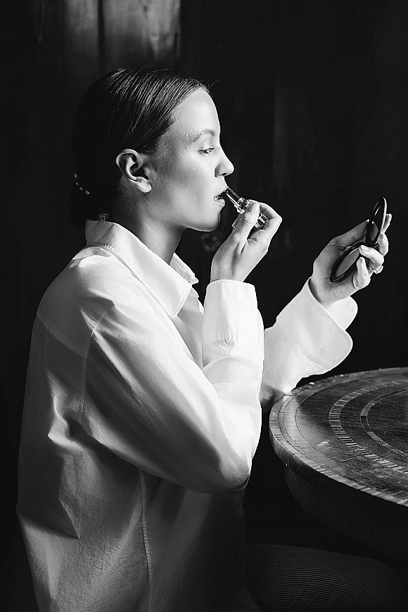 Bride getting ready, applying lipstick while holding a compact mirror, wearing a button-down shirt and stud earrings against a wood wall