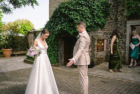 Wedding first look as bride in a satin gown with veil and bouquet reacts to groom in beige suit in an ivy stone courtyard doorway