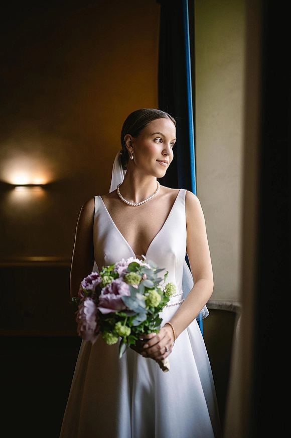 Bridal portrait of a bride holding bouquet in soft window light, wearing a veil and pearl necklace beside a neutral wall sconce
