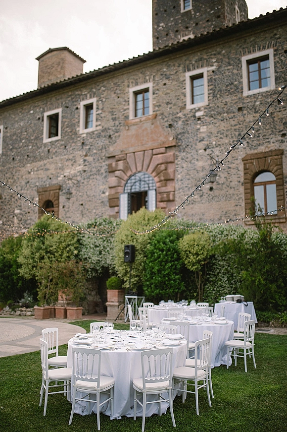 Outdoor reception setup with round wedding tables in white tablecloths, place settings and string lights beside a stone facade courtyard