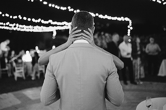 First dance wedding first dance moment with bride’s manicured hands and rings on groom’s jacket under string lights as guests watch outdoors at night