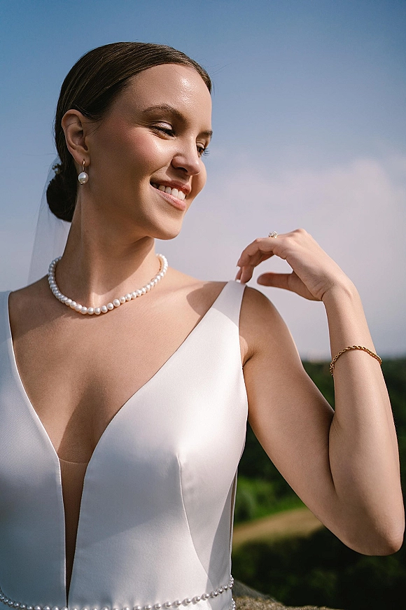 Bridal portrait of a modern bride in a minimalist white dress with pearl necklace and veil, looking away under blue sky and clouds