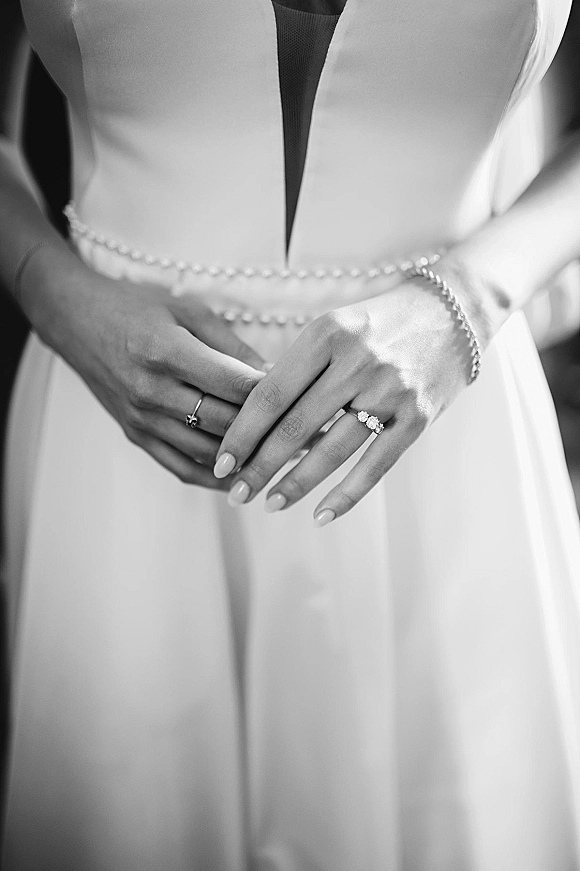 Wedding dress details with deep v wedding dress neckline and beaded waist belt, bride’s manicured hands showing engagement ring and band in soft indoor blur