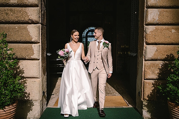 Wedding couple portrait of newlyweds holding hands, bride in veil with pink peony bouquet and groom in beige suit at stone doorway entrance