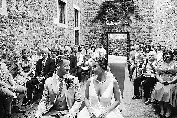 Ceremony moment at an outdoor wedding ceremony with bride and groom seated, smiling as guests watch in a stone courtyard aisle