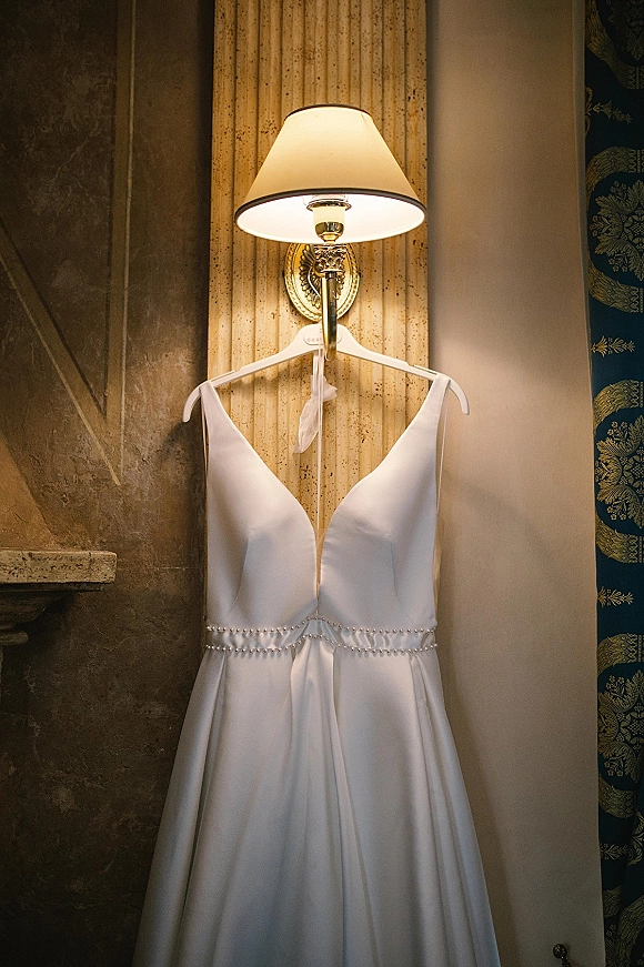 Wedding dress hanging on a white hanger, showing a v neck wedding dress bodice with pearl-beaded waist, beside a wall sconce on textured wallpaper