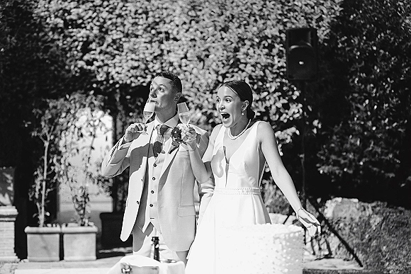 Wedding toast with bride and groom raising champagne flutes beside a white cake at an outdoor garden reception, greenery behind them
