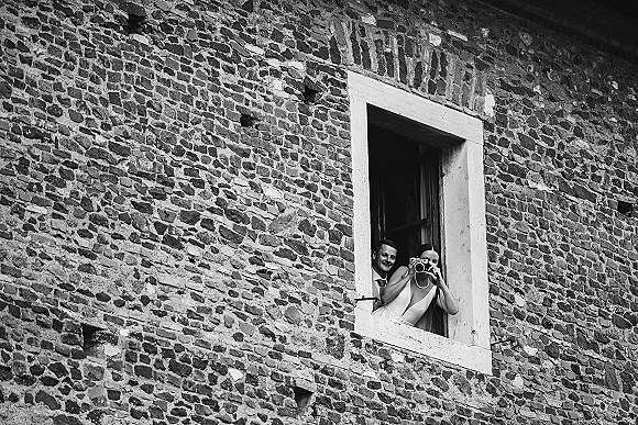 Couple portrait of bride with camera as groom peeks behind her, framed by a window in a stone wall, in black and white