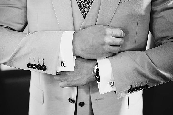 Groom suit details with wedding cufflinks on shirt cuffs, light suit jacket and patterned tie with wristwatch in an indoor setting