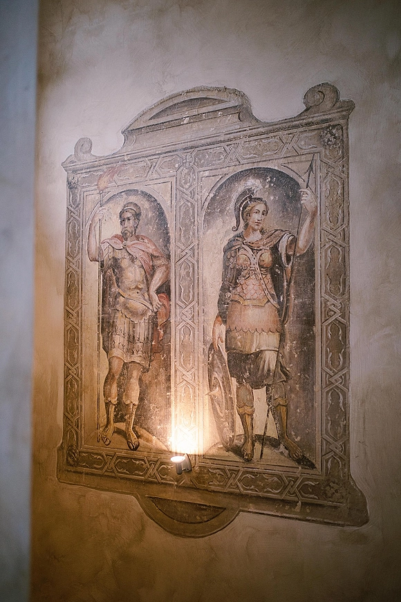 Wedding venue wall art featuring a fresco wall mural in a decorative frame, lit by a warm wall sconce against a textured plaster wall