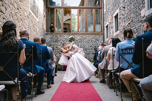 Wedding kiss as groom dips bride holding a bouquet, veil flowing on a red carpet aisle in a stone courtyard with guests and musicians