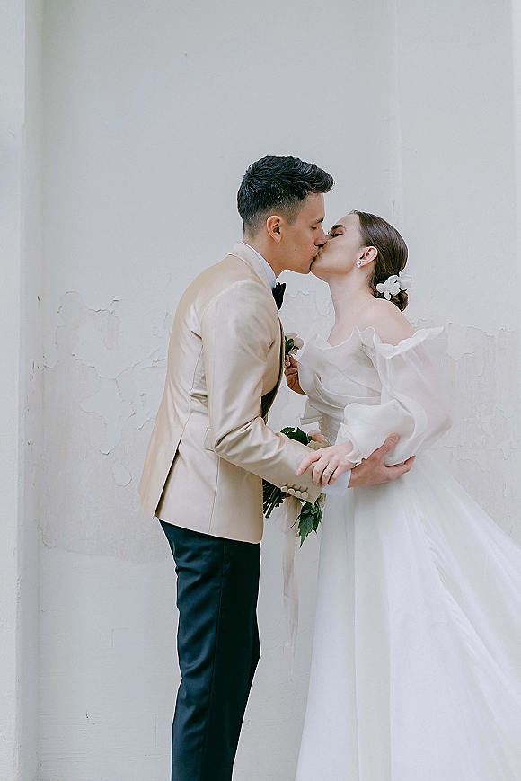 Wedding kiss portrait of bride and groom kissing in side profile, her off-shoulder dress and bouquet beside his beige suit against a white wall