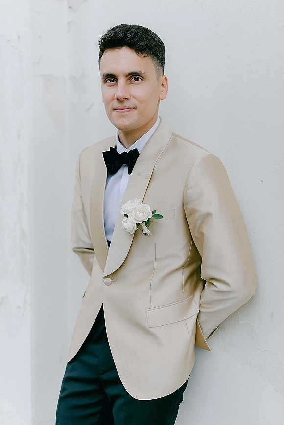Groom portrait in a tan tuxedo jacket with black bow tie and white rose boutonniere, hands in pockets against a white wall