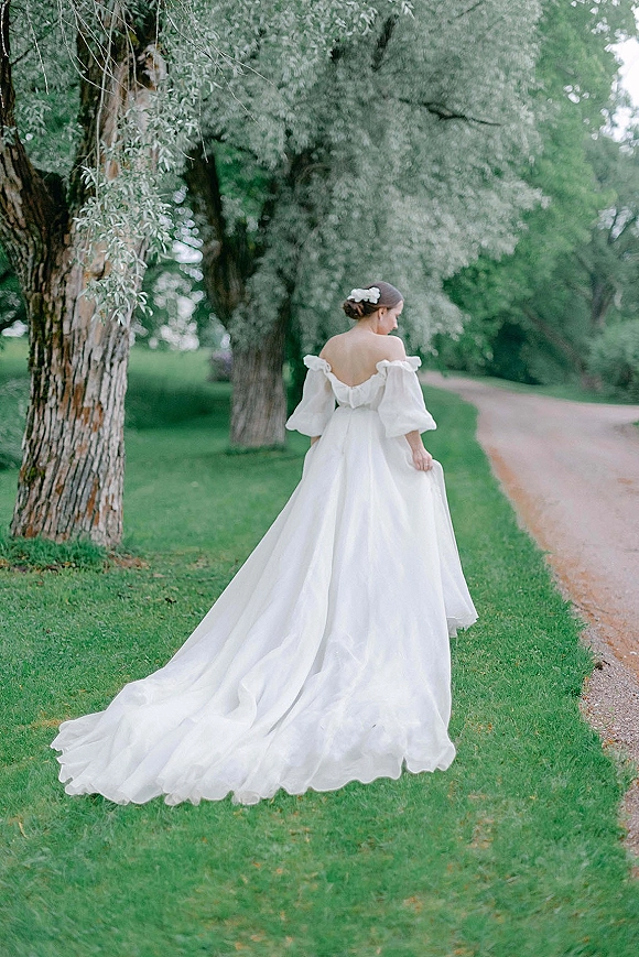 Bridal portrait of a bride in a backless wedding dress with off-the-shoulder sleeves and long train, walking along a garden path under trees