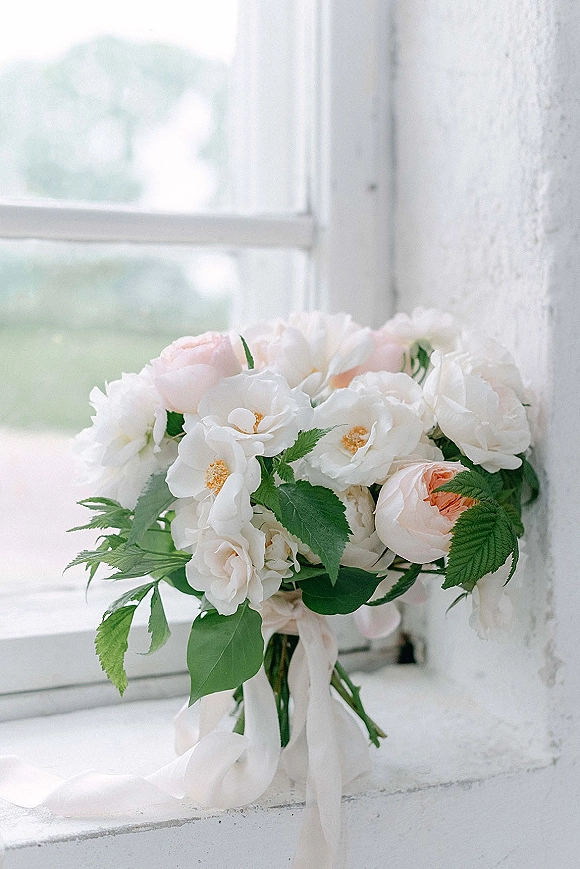 Bridal bouquet of white and blush flowers with greenery and a white ribbon wrap resting on a windowsill by a white wall