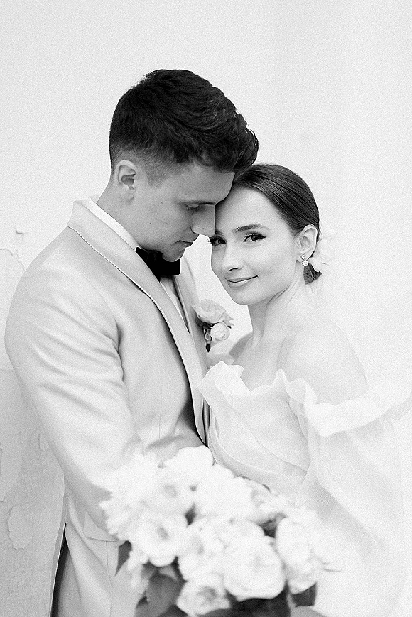 Couple portrait in a black and white wedding portrait as the groom kisses the bride’s forehead, her veil and bouquet against a plain wall