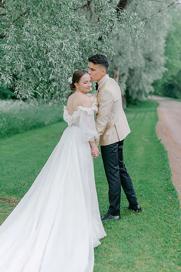 Couple portrait of groom kissing bride’s forehead, holding hands on a green lawn by a garden path, her off-shoulder gown and updo with hair flower