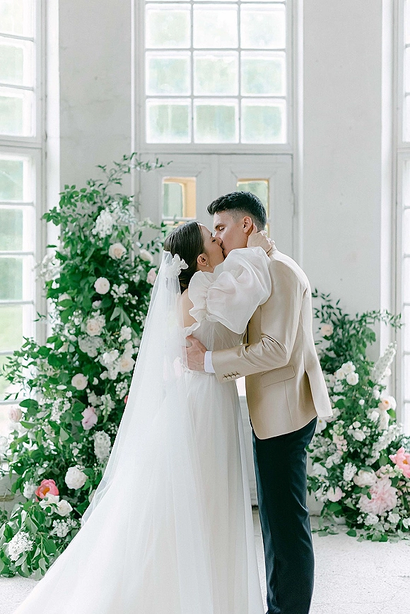 Wedding kiss portrait of bride and groom kiss by large windows, bride in puff sleeve gown and veil, holding his face amid florals