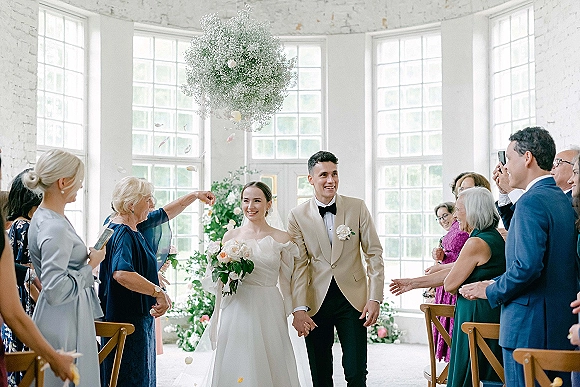 Wedding recessional as bride and groom walking aisle hand in hand while guests toss petals under hanging florals in a bright white brick venue