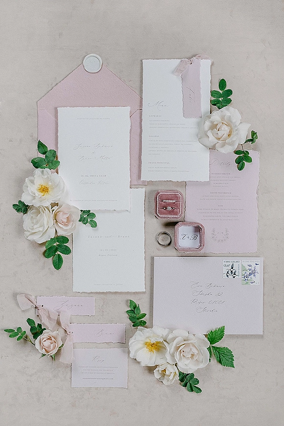 Invitation suite wedding invitation flat lay with deckle edge paper, blush envelope, wax seal and silk ribbon on neutral fabric with rings, roses and greenery