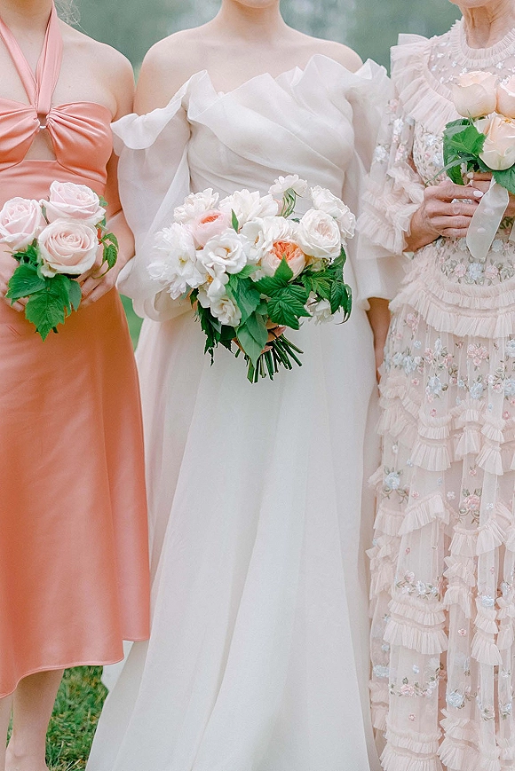 Bridesmaids with bouquets posing together, holding rose and greenery arrangements with ribbon, in satin and floral dresses on grass outdoors