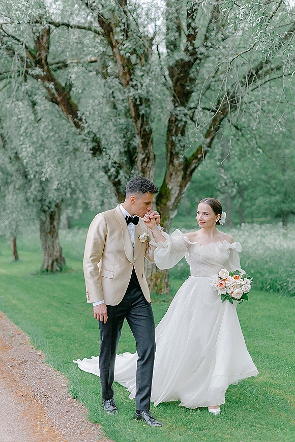 Couple portrait of groom kissing the bride’s hand as they walk on a garden path, bride in puff-sleeve gown holding a bouquet